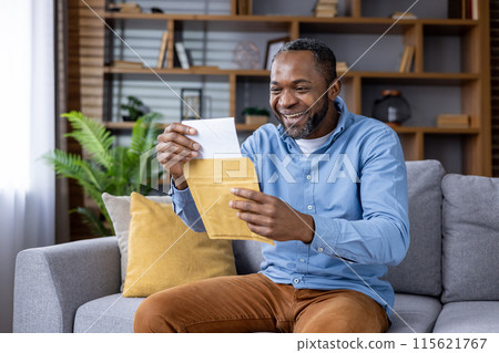 Joyful man wearing casual clothes and smiling while opening a letter on a sofa in a cozy living room. 115621767