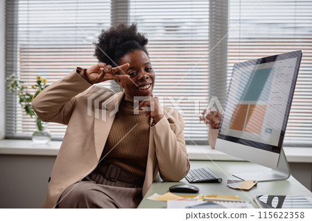 Medium shot portrait of happy optimistic Black woman doing victory gesture and looking smiling to camera, showing peace, v-sign with double fingers sitting against window in light office, copy space 115622358