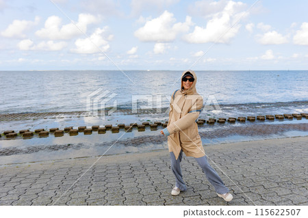 Smiling happy woman walking by Nord sea coast wear beige coat  sunglasses enjoys windy day cloudy sky background. Female person have fun hiking adventure travel at Baltic sea seaside 115622507