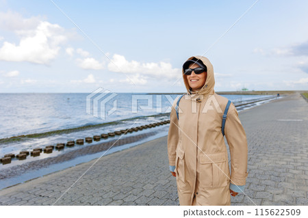 Smiling happy woman walking by Nord sea coast wear beige coat  sunglasses enjoys windy day cloudy sky background. Female person have fun hiking adventure travel at Baltic sea seaside 115622509