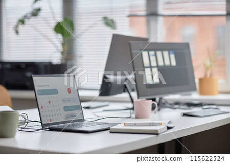 Side view shot in pastel colors of office workplace table with laptop displaying calendar page, notebook and computer monitors in background, focus on screen, copy space 115622524