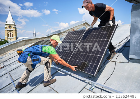 Workers building solar panel system on metal rooftop in urban setting. Two men installers carrying photovoltaic solar module outdoors. Alternative, green and renewable energy generation concept. Workers building solar panel system on metal rooftop in urban setting. Two men installers carrying photovoltaic solar module outdoors. Alternative, green and renewable energy generation concept. 115624495
