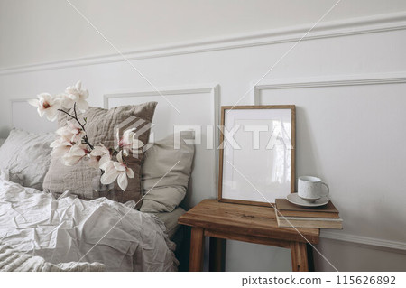 Blank wooden picture frame mockup on old book. Wooden night stand with fluted glass vase. Blooming magnolia tree branches. Scandinavian interior. Elegant bedroom. White wall background, stucco decor. 115626892