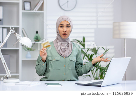 Confused woman in hijab holding credit card while using laptop in modern office setting. Confused woman in hijab holding credit card while using laptop in modern office setting. 115626929