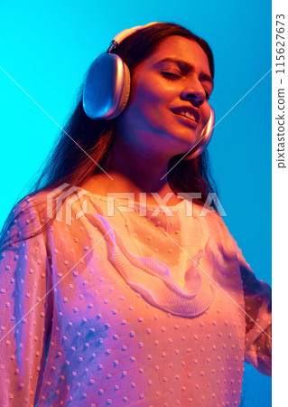 Portrait of young Indian woman listening music in in headphones, dancing and singing in neon light against blue gradient background. Portrait of young Indian woman listening music in in headphones, dancing and singing in neon light against blue gradient background. 115627673