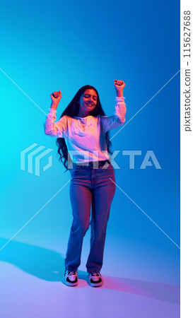 Dynamic portrait of young Indian woman dressed stylish clothes dancing in motion in neon light against blue gradient background. 115627688