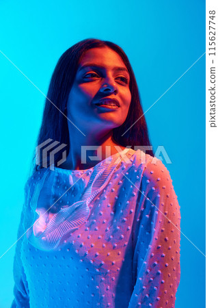 Portrait of young Indian woman dressed white festive shirt looking in distance in neon light against blue gradient background. Portrait of young Indian woman dressed white festive shirt looking in distance in neon light against blue gradient background. 115627748