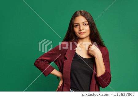 Professional portrait of Indian woman in burgundy blazer and black shirt, standing confidently looking at camera against green studio background. Professional portrait of Indian woman in burgundy blazer and black shirt, standing confidently looking at camera against green studio background. 115627803