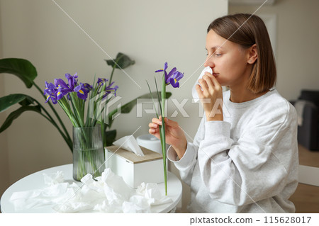 Allergic young woman holds iris flower, covers nose with paper tissue has runny nose, sneezes from flowers pollen at kitchen home by table. Girl with flu, itching or cough, seasonal allergy, rhinitis Allergic young woman holds iris flower, covers nose with paper tissue has runny nose, sneezes from flowers pollen at kitchen home by table. Girl with flu, itching or cough, seasonal allergy, rhinitis 115628017