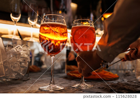 Bartender makes two glasses of cocktail Aperol spritz on bar counter, adds fresh orange slices. Typical alcoholic Italian beverage, aperitif made with Prosecco sparkling white wine and ice cubes. Bartender makes two glasses of cocktail Aperol spritz on bar counter, adds fresh orange slices. Typical alcoholic Italian beverage, aperitif made with Prosecco sparkling white wine and ice cubes. 115628028