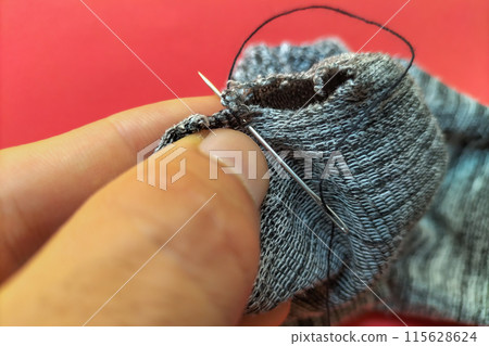 Hands of a white man sewing a hole in a striped gray sock with a sewing needle and black thread against a red background. Repair of clothes and ammunition, survival lifestyle, autonomy. 115628624