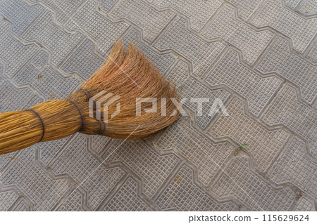 A brown natural broom made of plants sweeps the textured pavement paved with cobblestones. Maintaining the cleanliness of the house space, entrances and courtyard. 115629624