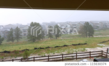 A vivid image capturing a heavy hailstorm over a suburban neighborhood. The storm pelts down on houses, trees, and an open field, creating a dramatic scene of intense weather conditions. A vivid image capturing a heavy hailstorm over a suburban neighborhood. The storm pelts down on houses, trees, and an open field, creating a dramatic scene of intense weather conditions. 115630374