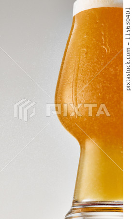 Close-up of shaped glass filled with a golden, carbonated beer with thick, creamy foam cascades over rim against grey studio background. Close-up of shaped glass filled with a golden, carbonated beer with thick, creamy foam cascades over rim against grey studio background. 115630401