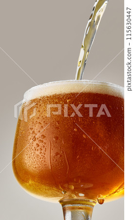 Close-up of goblet filled with rich, amber, craft beer being poured into glass against grey studio background. Close-up of goblet filled with rich, amber, craft beer being poured into glass against grey studio background. 115630447