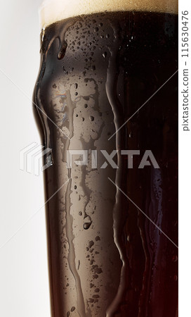 Close-up of pint glass filled with dark, cold, foamy craft beer with droplets on condensation on glass against grey studio background. Close-up of pint glass filled with dark, cold, foamy craft beer with droplets on condensation on glass against grey studio background. 115630476