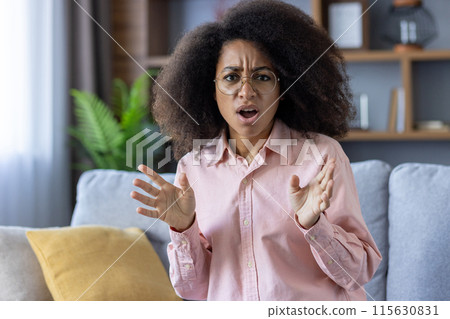 Surprised woman with curly hair and glasses reacting with open hands while sitting on a couch at home 115630831