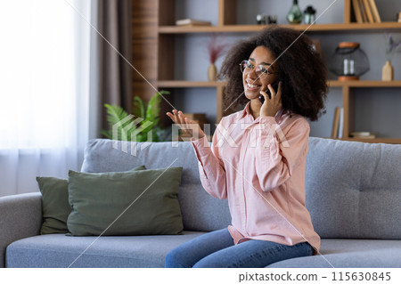 Smiling woman with curly hair having a pleasant phone conversation while sitting on a couch in a cozy living room. Smiling woman with curly hair having a pleasant phone conversation while sitting on a couch in a cozy living room. 115630845