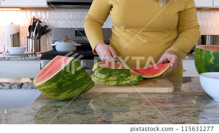 A woman in a yellow sweater slices a fresh watermelon on a cutting board in a modern kitchen. The kitchen features white cabinetry, a hexagonal tile backsplash, and various cooking utensils on the 115631277