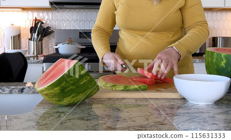 A woman in a yellow sweater slices a fresh watermelon on a cutting board in a modern kitchen. The kitchen features white cabinetry, a hexagonal tile backsplash, and various cooking utensils on the A woman in a yellow sweater slices a fresh watermelon on a cutting board in a modern kitchen. The kitchen features white cabinetry, a hexagonal tile backsplash, and various cooking utensils on the 115631333