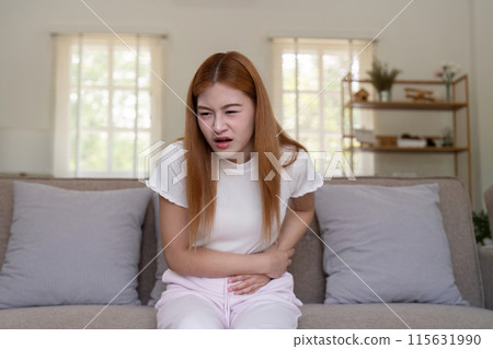 Young Woman Experiencing Stomach Pain While Sitting on a Couch in a Bright Living Room 115631990