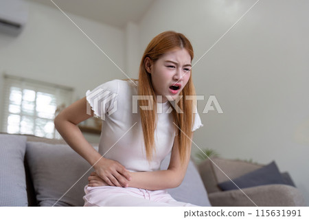 Woman Experiencing Stomach Pain While Sitting on Couch, Holding Her Abdomen in Discomfort 115631991