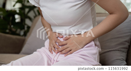 Woman Experiencing Stomach Pain While Sitting on a Couch in a Home Setting, Clutching Her Abdomen in Discomfort 115631992