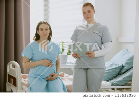 Pregnant woman sitting on hospital bed and holding her belly lovely while nurse standing next to her and holding medical chart 115632056