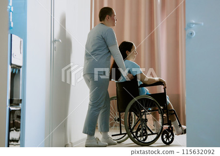 Nurse dressed in light gray scrubs holding handles of wheelchair and her pregnant patient resting while sitting in it 115632092
