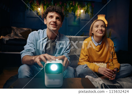 Man and a woman are sitting on a couch watching a movie on a projector 115632424