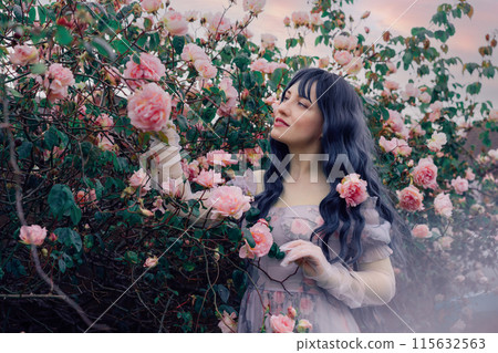 Fairy tale woman portrait. Young beautiful woman Fairy holds pink flower rose on blooming bush background. Hairstyle loose green hair, romantic purple medieval vintage dress. Summer fantasy garden. 115632563
