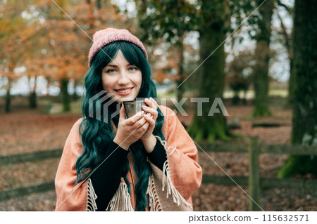 Smiling Woman in hat wrapped in Shawl Poncho drinking hot drink from thermos cup during autumn walk in forest with fall leaves. Feeling harmony, relax, personal fulfillment. Local travel lifestyle. 115632571