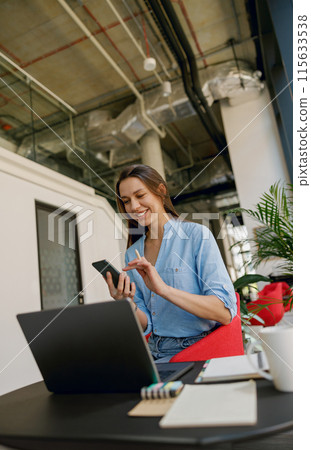 A young woman is happily working remotely from a modern office space, using a laptop and phone 115633538