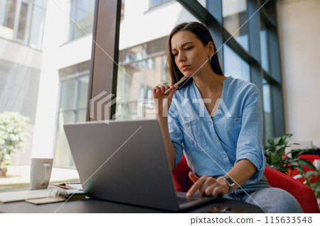 A professional woman is focusing on work at a laptop in a modern office with large windows 115633548
