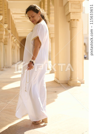 Elegant woman in white dress strolling in sunlit architectural corridor Elegant woman in white dress strolling in sunlit architectural corridor 115633651