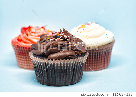 Three cupcakes with colorful sprinkles standing on blue background Three cupcakes with colorful sprinkles standing on blue background 115634155