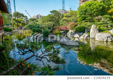 Amazing landscape in Japanese garden of Monaco with pond , large rocks (in water) and garden path.Red Japanese bridge in distance Amazing landscape in Japanese garden of Monaco with pond , large rocks (in water) and garden path.Red Japanese bridge in distance 115634235