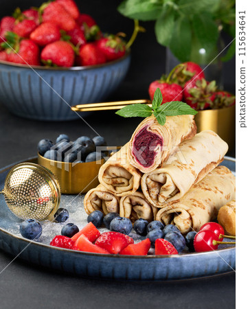 Crepes stacked in a pile, one of which is cut and filled with berry filling. The plate is also decorated with blueberries, strawberries Crepes stacked in a pile, one of which is cut and filled with berry filling. The plate is also decorated with blueberries, strawberries 115634641