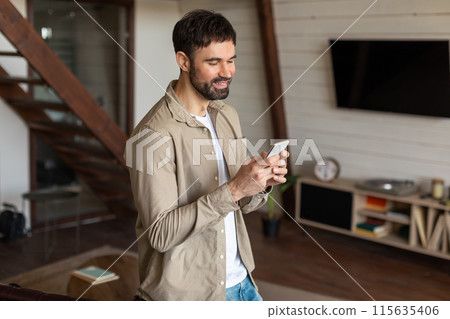 A man is standing in the living room, fixated on his cell phone screen. The room is furnished with a sofa, coffee table, and TV. The man appears focused on the device in his hands 115635406