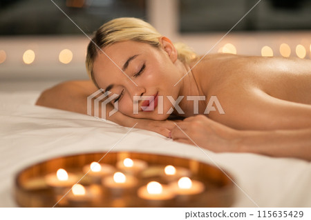 A woman is lying on a bed, with flickering candles placed around her at SPA. The room is dimly lit, creating a warm and intimate atmosphere. A woman is lying on a bed, with flickering candles placed around her at SPA. The room is dimly lit, creating a warm and intimate atmosphere. 115635429