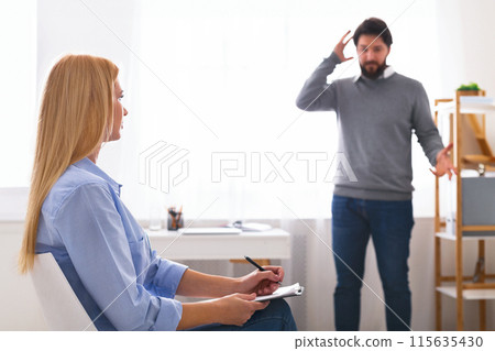 A man stands gesturing with his hand to his head in a sign of distress or making a point during a therapy session, as woman counselor, seated across from him 115635430