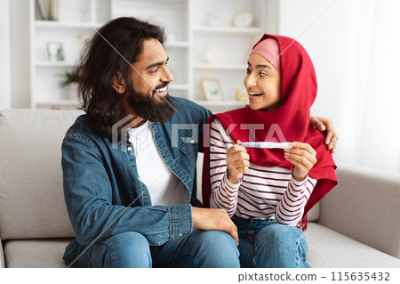 Happy Muslim Couple Holding Positive Pregnancy Test Sitting On Couch At Home. Young Arab Spouses Awaiting Baby. Childbirth And Family Life Moments. We'll Be Parents Concept 115635432