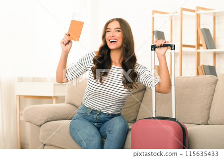 A smiling young woman holds up her passport and boarding passes excitedly as she sits on a wheeled suitcase, prepared to embark on a journey 115635433