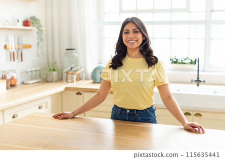 A middle eastern woman with a yellow shirt is standing at a kitchen counter, engaging in household activities. She appears focused and busy as she prepares food or cleans the kitchen. 115635441