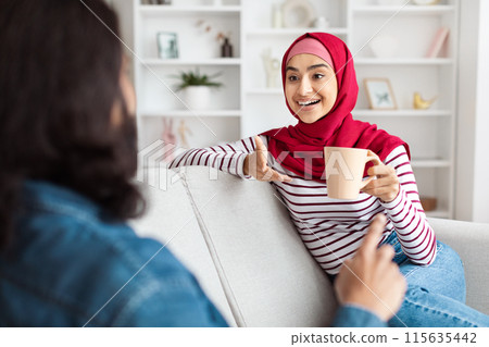 A joyful eastern woman dressed in a red hijab and striped top relaxes on a white couch, gesturing animatedly with a mug in her hand as she engages in a lively chat with her husband A joyful eastern woman dressed in a red hijab and striped top relaxes on a white couch, gesturing animatedly with a mug in her hand as she engages in a lively chat with her husband 115635442