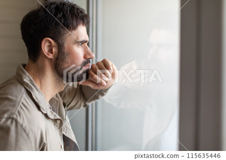 A man stands near a window, gazing outside with his hand resting on his face. He appears contemplative and deep in thought as he observes the view before him. 115635446