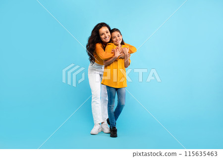 Happy european mother hugging her lovely daughter, standing over blue studio background, full length shot, free space. Young mom and he child girl bonding and smiling to camera Happy european mother hugging her lovely daughter, standing over blue studio background, full length shot, free space. Young mom and he child girl bonding and smiling to camera 115635463