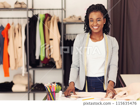 Cheerful African American lady fashion designer posing with measuring tape in studio, surrounded by garments 115635476