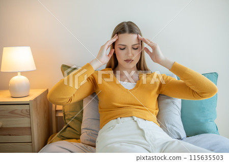 A girl is seated on a bed, holding her head with her hands. She appears deep in thought or experiencing distress. Her body language suggests emotional turmoil or physical discomfort. A girl is seated on a bed, holding her head with her hands. She appears deep in thought or experiencing distress. Her body language suggests emotional turmoil or physical discomfort. 115635503