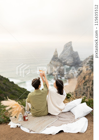 Back view of romantic couple celebrating their engagement or honeymoon on the coastline, drinking champagne and clinking glasses, enjoying ocean shore, copy space, vertical shot 115635512